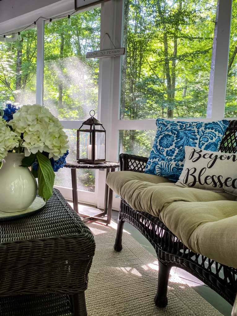 view of hydrangeas, lit candle in Iron Lantern Tabletop Lantern and Emilie Indoor / Outdoor Throw Pillow Cover & Insert on wicker couch.