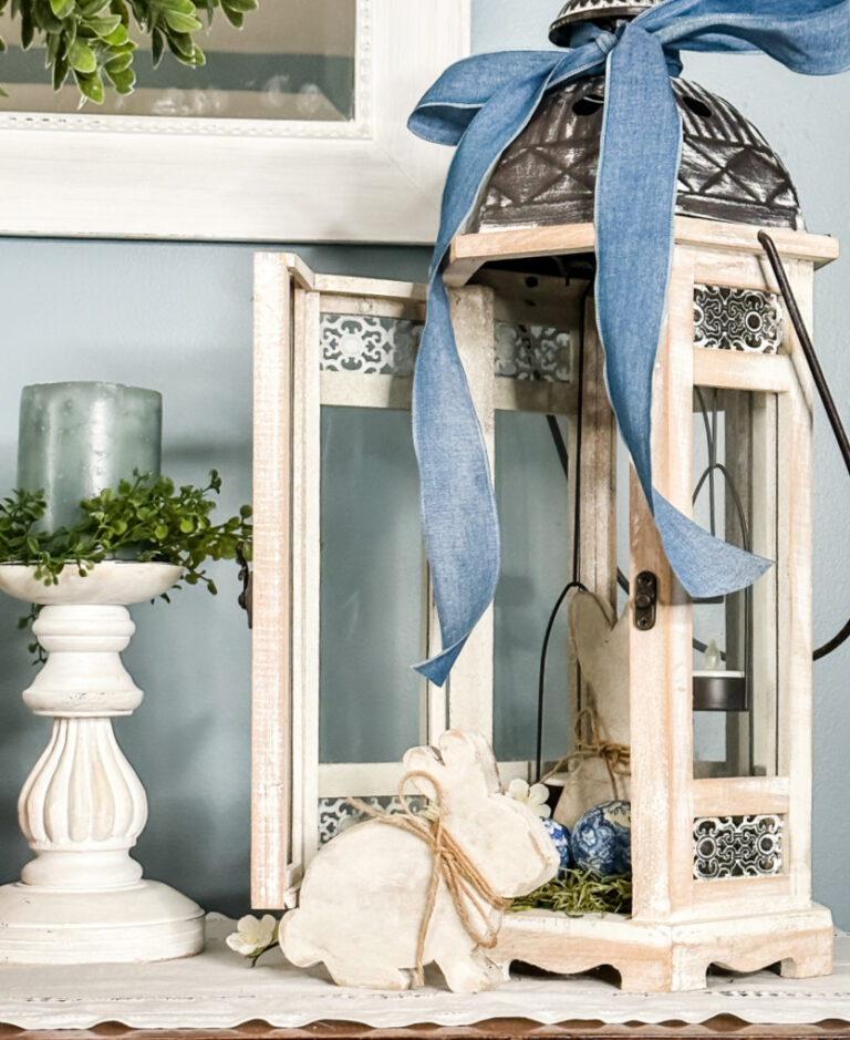 whitewashed lantern on a side table decorated with wooden Easter bunnies and a blue ribbon
