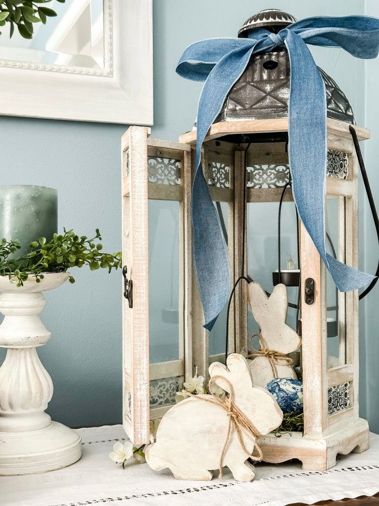 whitewashed lantern on a side table decorated with wooden Easter bunnies, moss, faux greens, decoupaged eggs, and a blue ribbon