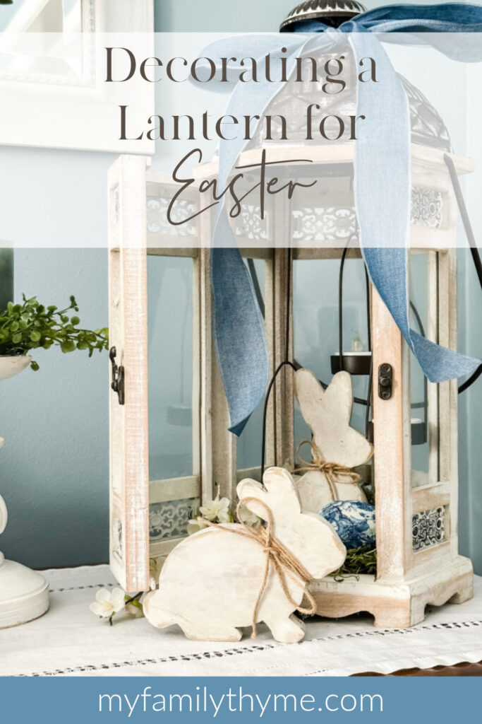 whitewashed lantern on a side table decorated with wooden Easter bunnies, moss, faux greens, decoupaged eggs, and a blue ribbon