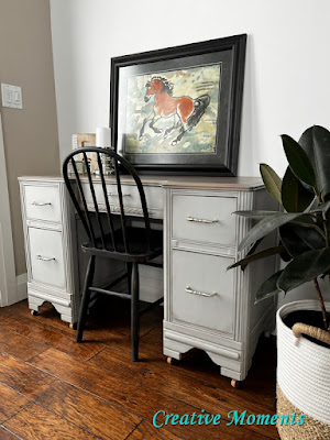 desk or vanity painted light gray with a black chair and a painting leaning on the desk top