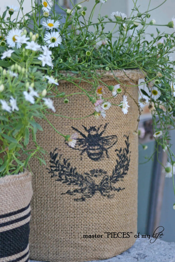 diy burlap covered garden container with black bee on it containing white flowering plant in it.