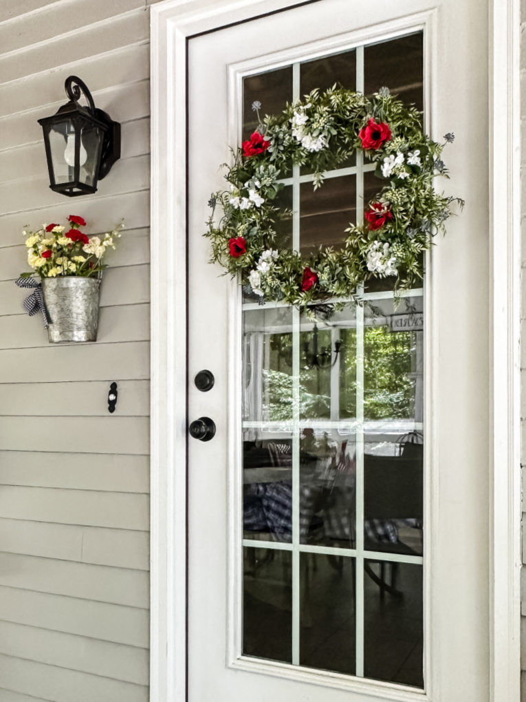view of white back door with red, white, and blue floral wreath and galvanized bucket with flowers on wall of house