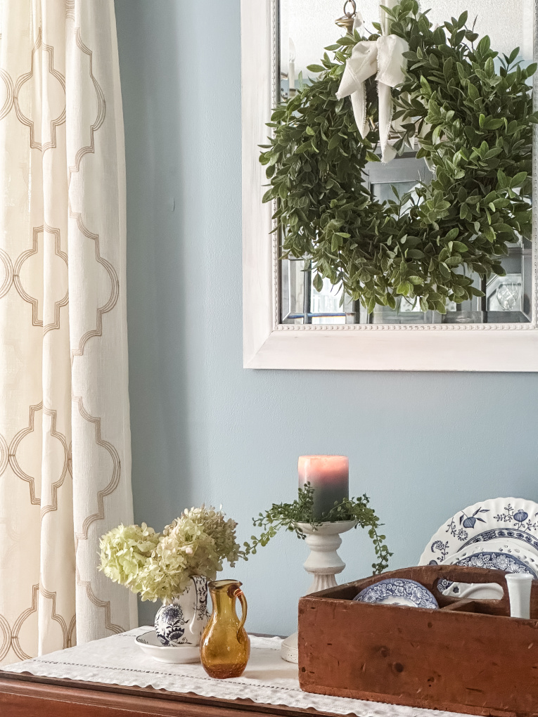 early fall decorating traditional dining room with blue painted walls and side table with candles and dried hydrangeas. White framed mirror with green wreath hanging.