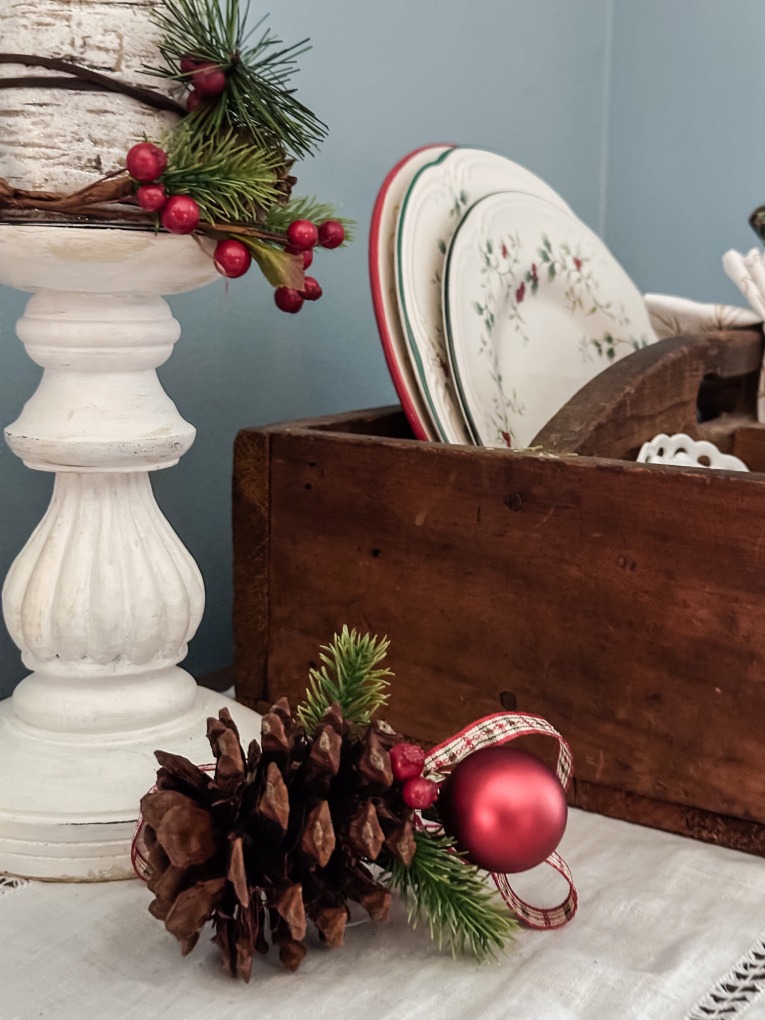 close up of pine cone ornament on side table, painted candlestick and Christmas dishes in vintage tool box.