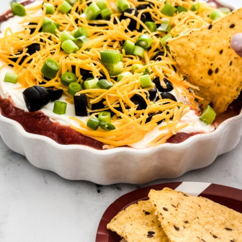 Mexican Layer Dip in a white round dish with a tortilla chip and chips on a football plate