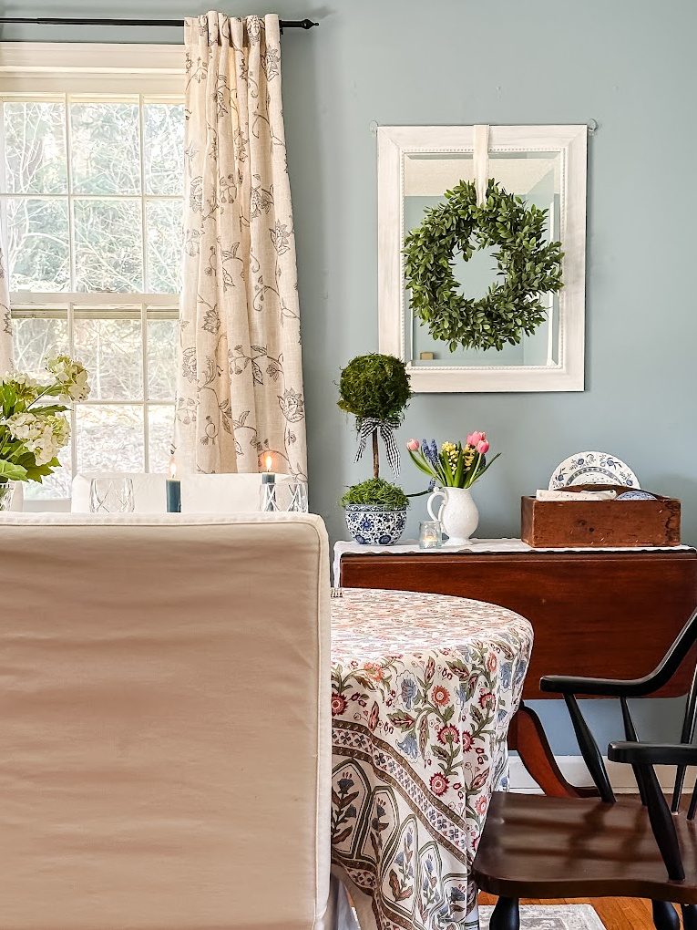 blue dining room with white mirror with wreath on it. Side table with topiary and spring flowers