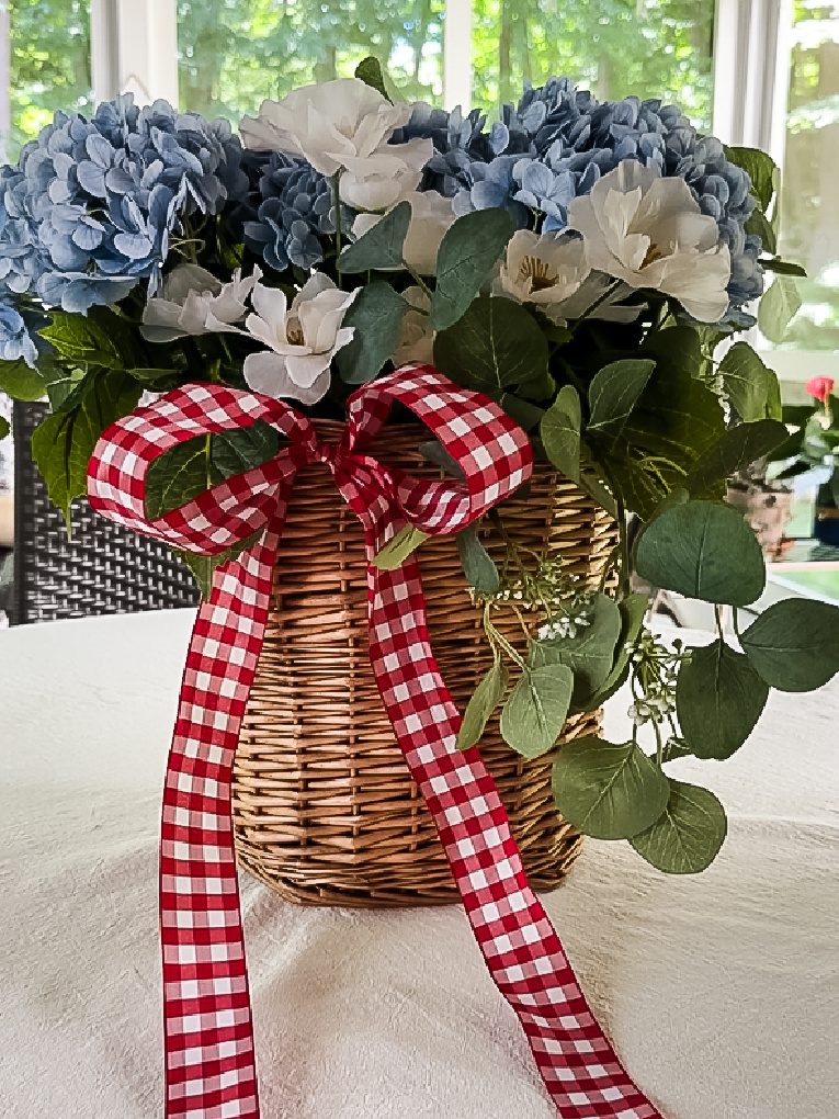 wicker door basket filled with blue and white flowers, greenery, and red and white gingham ribbon