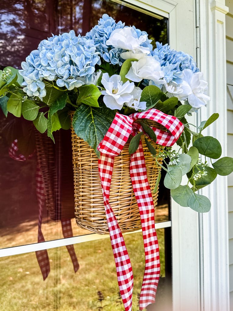wicker summer floral door basket filled with blue and white flowers, greenery, and red and white gingham ribbon