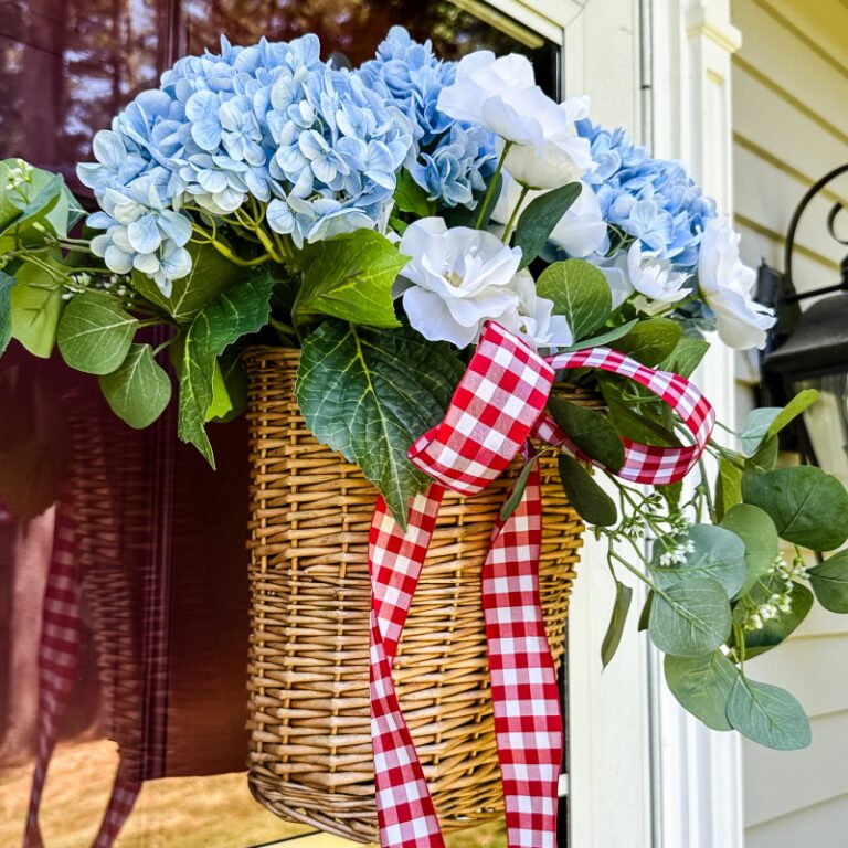 wicker door basket filled with blue and white flowers, greenery, and red and white gingham ribbon