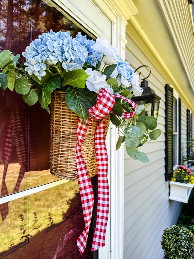 view of wicker door basket filled with blue and white flowers, greenery, and red and white gingham ribbon on door of grey house with window boxes