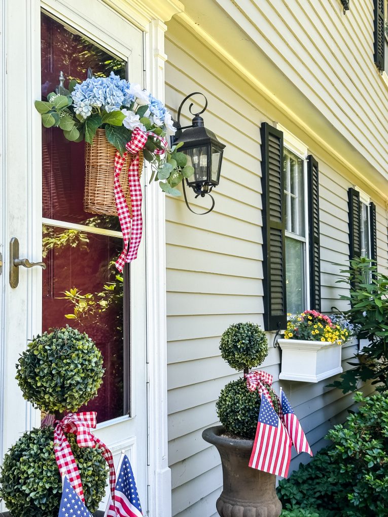 view of wicker door basket filled with blue and white flowers, greenery, and red and white gingham ribbon on door of grey house with topiaries and window boxes